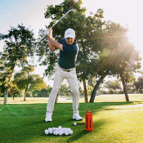A man in a golf swing on a sunny golf course, wearing a navy polo shirt, white pants, and a white cap. A pile of golf balls and an orange Ranger Turtlebox sit on the grass in the foreground, with trees and bright sunlight in the background.