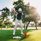 A man in a golf swing on a sunny golf course, wearing a navy polo shirt, white pants, and a white cap. A pile of golf balls and an orange Ranger Turtlebox sit on the grass in the foreground, with trees and bright sunlight in the background.