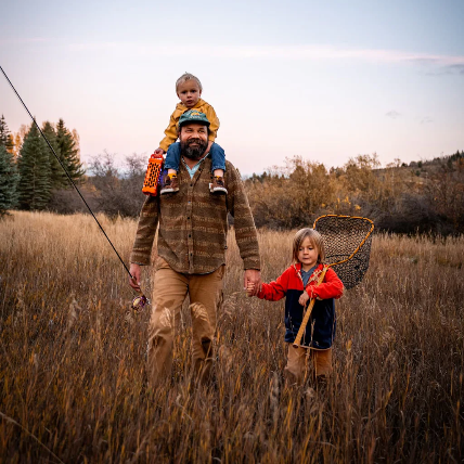 Man with two children in a field, holding a Turtlebox Ranger,  fishing gear against a clear sky.