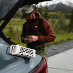 Person standing next to a car with an open trunk, holding a phone, and featuring a Turtlebox Ranger magnetically attached to a car in a natural setting.