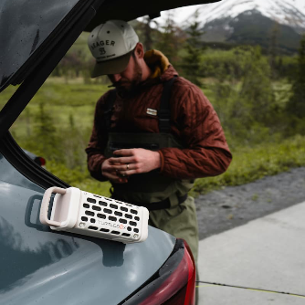 Person standing next to a car with an open trunk, holding a phone, and featuring a Turtlebox Ranger magnetically attached to a car in a natural setting.