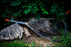 A black fishing net with a gray handle and orange accents resting on a weathered log in a forest setting with green foliage in the background.