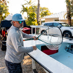 A man wearing a blue cap, sunglasses, and a gray hoodie stands beside a light blue boat, holding a large fishing net in an outdoor driveway with palm trees and parked cars in the background.