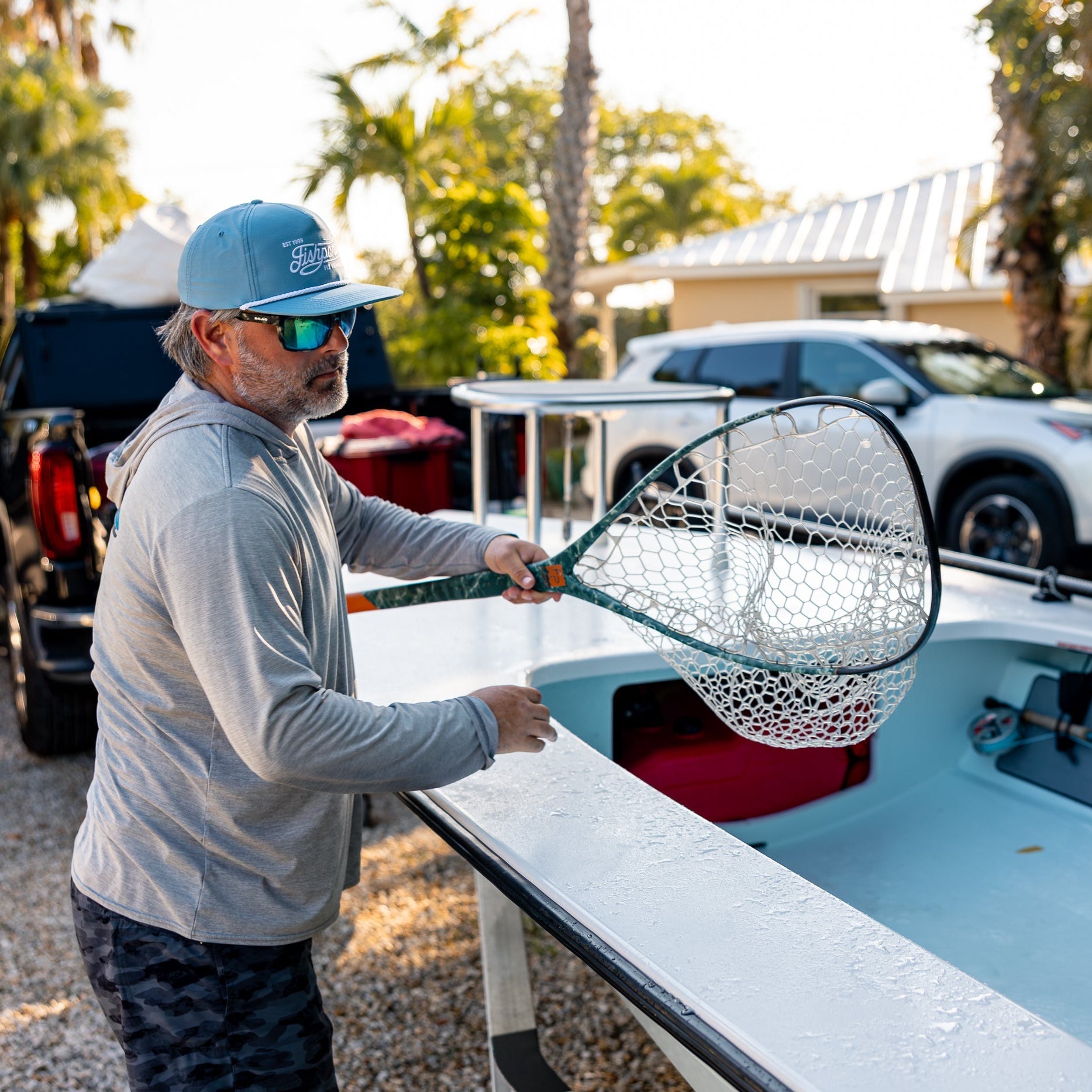 A man wearing a blue cap, sunglasses, and a gray hoodie stands beside a light blue boat, holding a large fishing net in an outdoor driveway with palm trees and parked cars in the background.