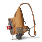 A brown tactical sling backpack shown from a side profile on a white background, featuring a side pocket holding red and black items, a small flap with yellow and white details, and a black circular attachment at the bottom.