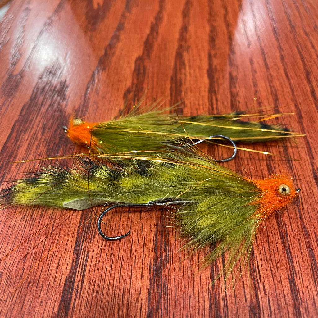 Two olive and orange artificial fishing flies designed for steelhead and salmon fishing, laid out on a wooden surface.
