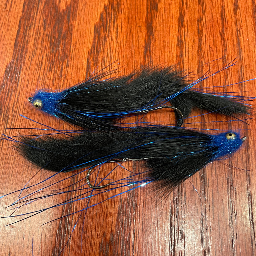 Two black and blue artificial fishing flies designed for steelhead and salmon fishing, laid out on a wooden surface.