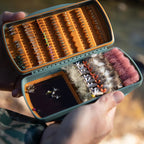 Open fly fishing box with various flies held by a person against a blurred natural background