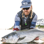Man holding a large striped bass by a body of water, wearing a YETI cap.