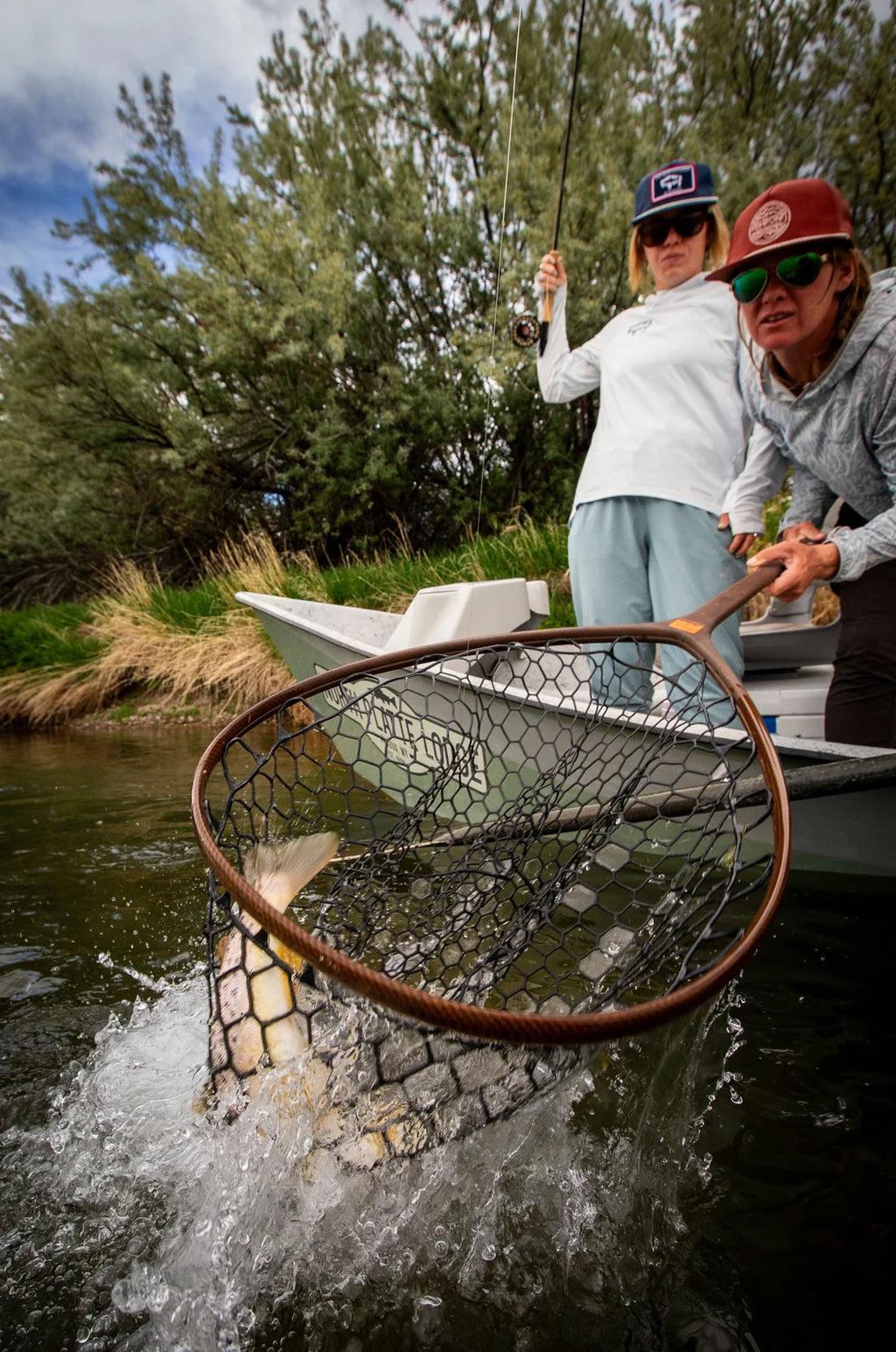 Two people in a small boat on a river or lake, with one person holding a large landing net and another holding a fishing rod. Trees and grass are visible in the background, and a fish is visible inside the net.