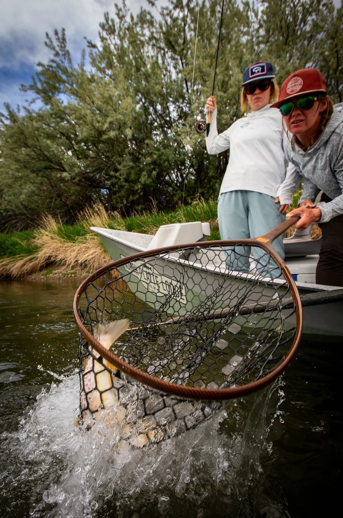 Two people in a small boat on a river or lake, with one person holding a large landing net and another holding a fishing rod. Trees and grass are visible in the background, and a fish is visible inside the net.