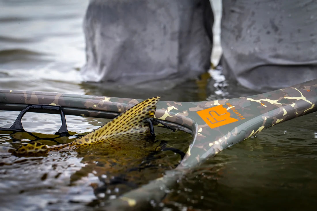 A close-up of a camouflage-patterned landing net resting in shallow water, with a bright orange sticker on the handle and blurred gray boots standing in the background.