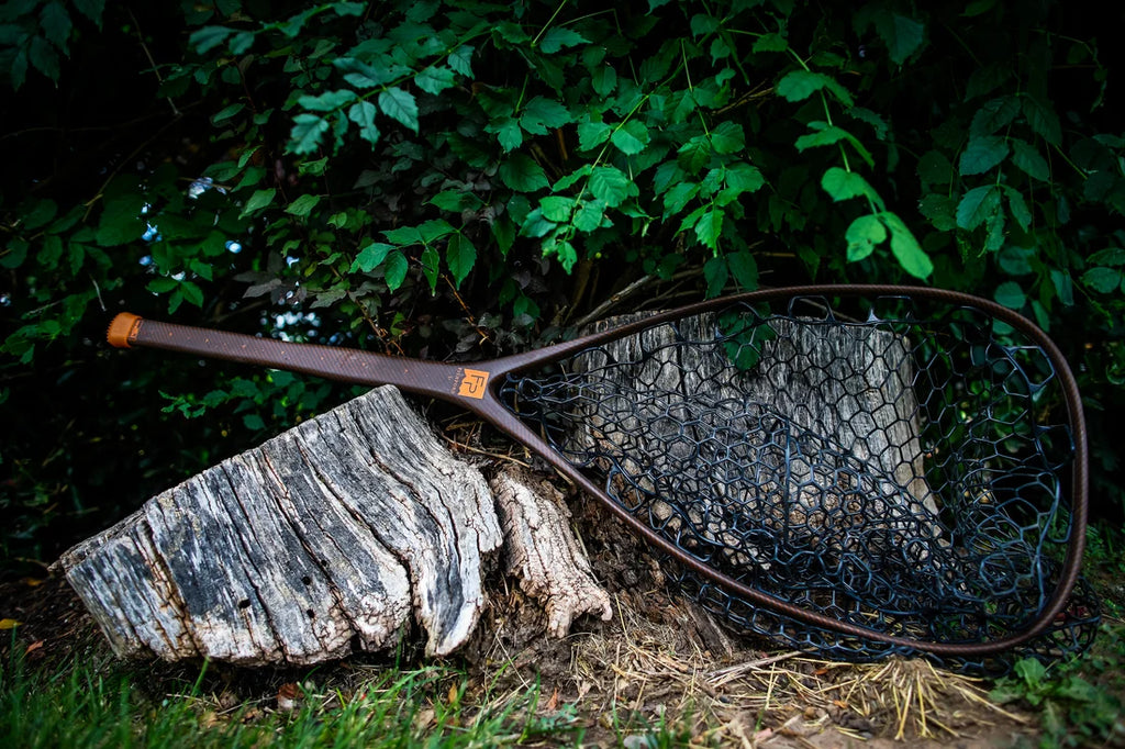 A Fishpond fishing net with a dark brown handle and black mesh lies resting on a weathered tree stump in an outdoor setting with green foliage in the background.