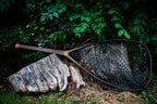 A Fishpond fishing net with a dark brown handle and black mesh lies resting on a weathered tree stump in an outdoor setting with green foliage in the background.