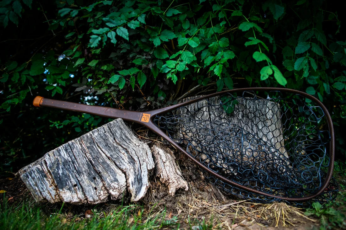 A Fishpond fishing net with a dark brown handle and black mesh lies resting on a weathered tree stump in an outdoor setting with green foliage in the background.