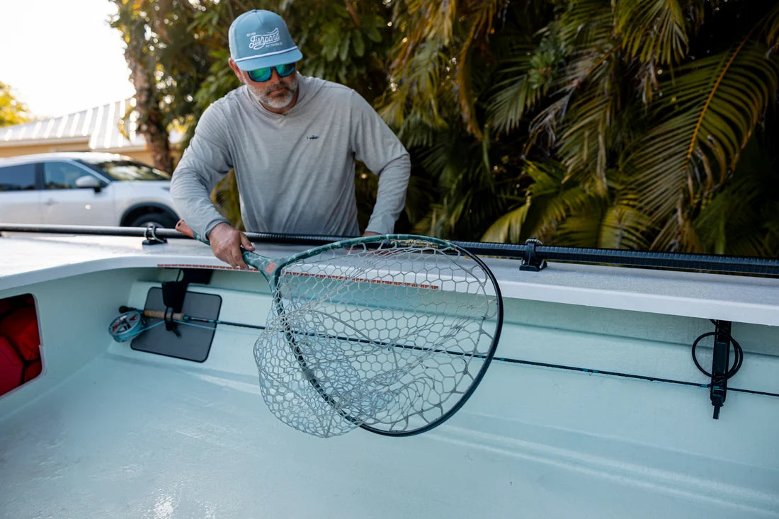 A man wearing a gray long-sleeve shirt and a blue cap stands in the bow of a boat, holding a large black-framed landing net with a gray mesh. Palm trees and a white car are visible in the background.