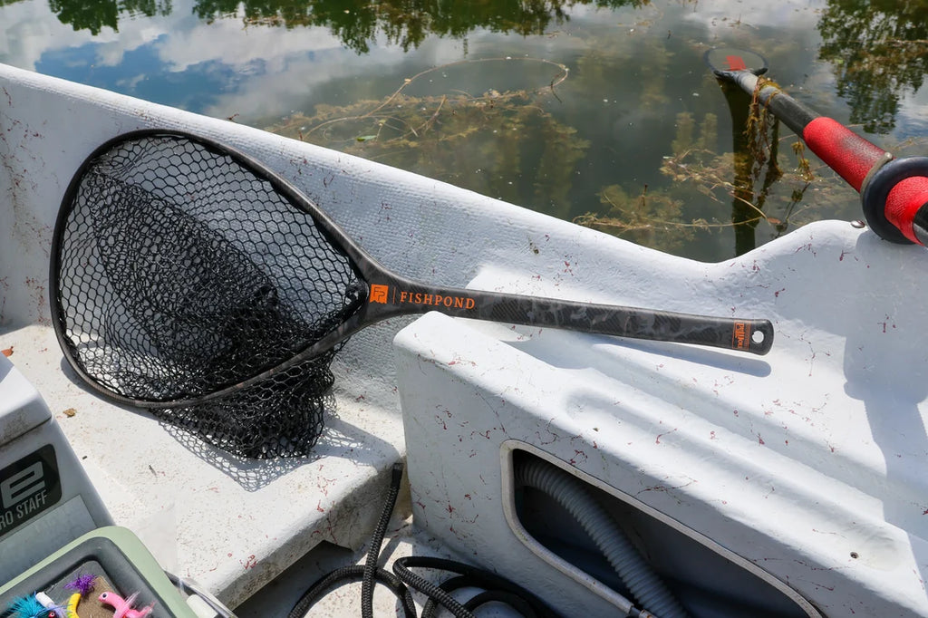 A black mesh fishing net with a black handle and orange 'FISHPOUND' logo, resting on the white gunwale of a boat next to water.