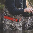 Person wearing camouflage waders and a red Simms Wading Belt - 2", standing in water and fishing.
