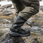 Person wearing black waterproof boots and green pants standing on a rocky surface with water.