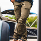 Person wearing brown Simms MEN's Rogue Softshell Pants and a blue shirt standing on a dock with a boat in the background.