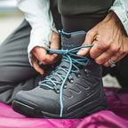 Person tying the laces of a black Simms WOMEN's Freestone Wading Boot - Felt Soles with teal laces on a pink surface.