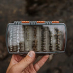 Hand holding a Fishpond fly fishing fly box with flies against a blurred natural background