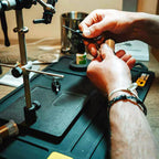 Person using a fly tying bobbin on a workbench with tools and equipment in the background