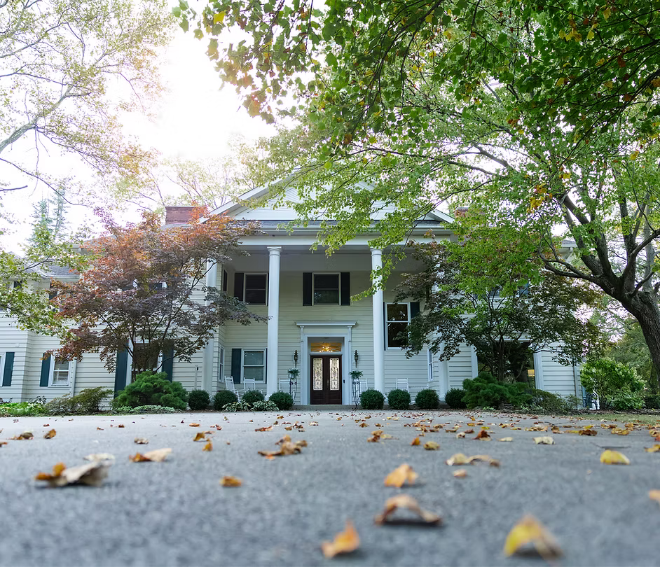 White colonial-style house with columns and a porch, surrounded by trees and fallen leaves.