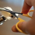 Person holding yellow-handled curved scissors near a metal object with a blurred background