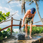 Man with a blue cap and camouflage shorts standing on a stone ledge with palm trees and a Turtlebox Original Gen 3 .