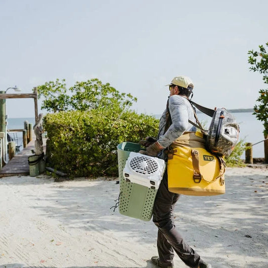 Person carrying a white Turtlebox Original Gen 3 speaker and green fishing chair by a dock with water and trees in the background.