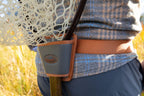 A person wearing a gray checkered shirt and a brown belt with a Fishpond net holster attached to it, holding a fishing net.