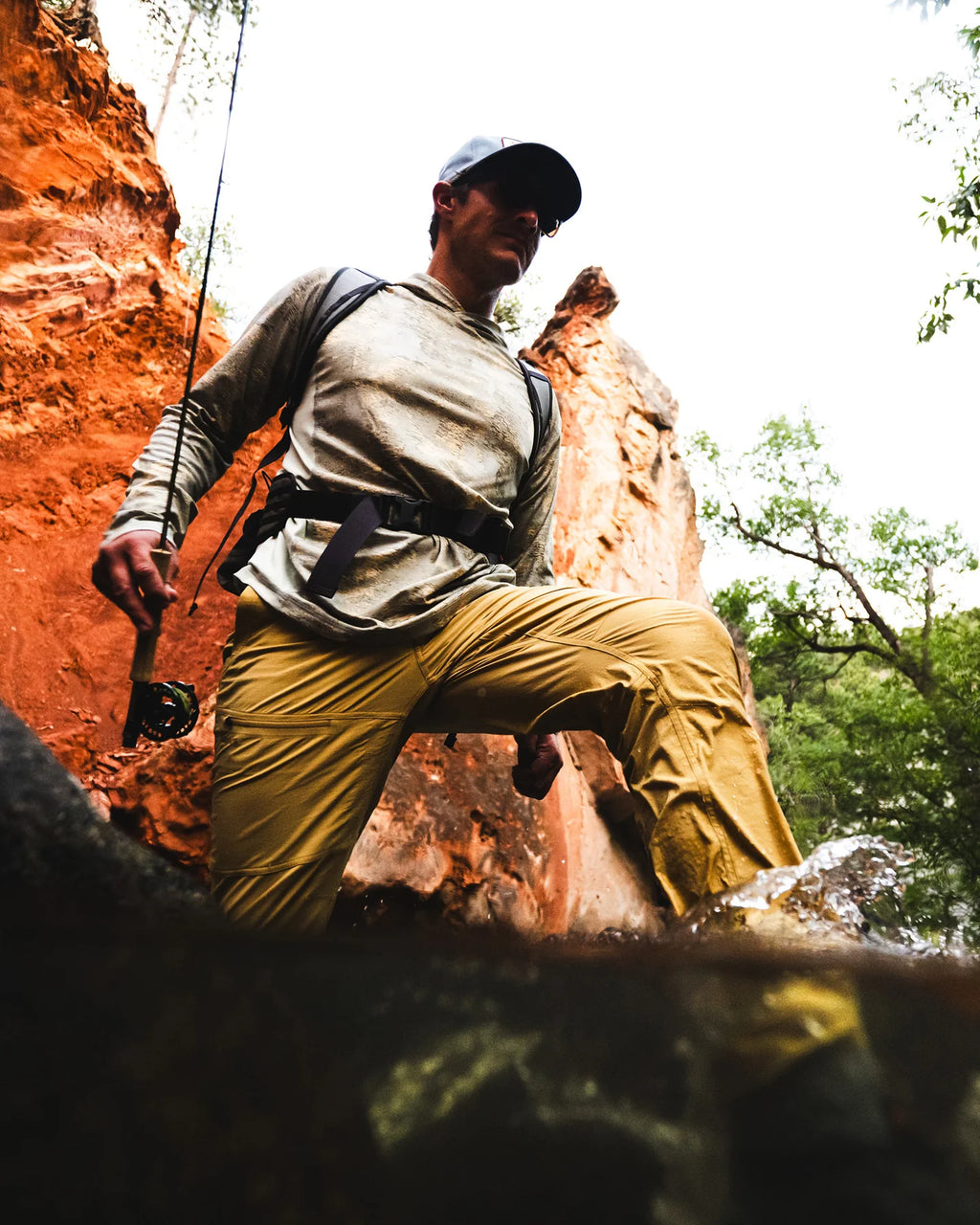 A man in outdoor fishing attire, including a gray long-sleeve shirt, tan pants, and a black cap, stands on a rock in a natural rocky environment holding a fishing rod.