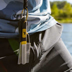 Person wearing a camouflage jacket with a fishing comb attached, standing by a body of water.