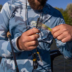 Person in a blue camouflage jacket holding a fishing lure and tools outdoors.