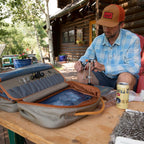 Man sitting at a table with fly tying bag and gear and a can of beer, near a log cabin.