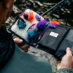 Person holding a black Fishing lure Yakoda STS Stash with colorful fishing lures inside, outdoors.