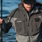 Man wearing a Simms jacket holding a fishing rod with water in the background