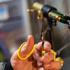 Hand holding yellow Loon Ergo All Purpose Scissors with a blurred background of a fly tying vise.
