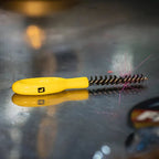A Loon Ergo Dubbing Brush with a yellow powder-coated handle and black nylon bristles, placed on a metal surface.