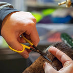 Person trimming a deer hair with yellow-handled scissors in a blurred indoor setting