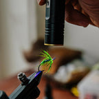 Close-up of a hand holding a black flashlight illuminating a green and blue fishing lure.