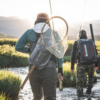 Two people walking through water with fishing gear, wearing backpacks with 'Simms' branding.