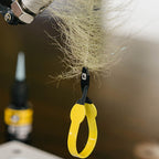 Close-up of a yellow Loon hackle plier tool with a black handle attached to a fishing fly, with a blurred background.