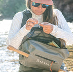 Person outdoors by a river, wearing a cap and sunglasses, with a Simms bag.