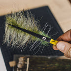 Hand holding a fly fishing hook with attached artificial grass skirt against a blurred background