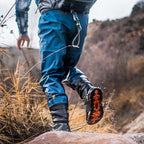 Person wearing blue hiking pants and Simms MEN's Freestone Wading Boot - Rubber Soles with orange sole stepping onto a rock in a natural setting.