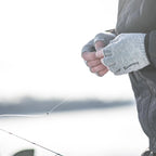 Person wearing gray gloves with 'Simms' branding, holding a fishing rod with a blurred water background.