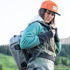 Person wearing a green jacket, orange cap, and gray backpack in a mountainous area.