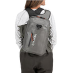 Person wearing a gray backpack with Simms logo, standing against a white background